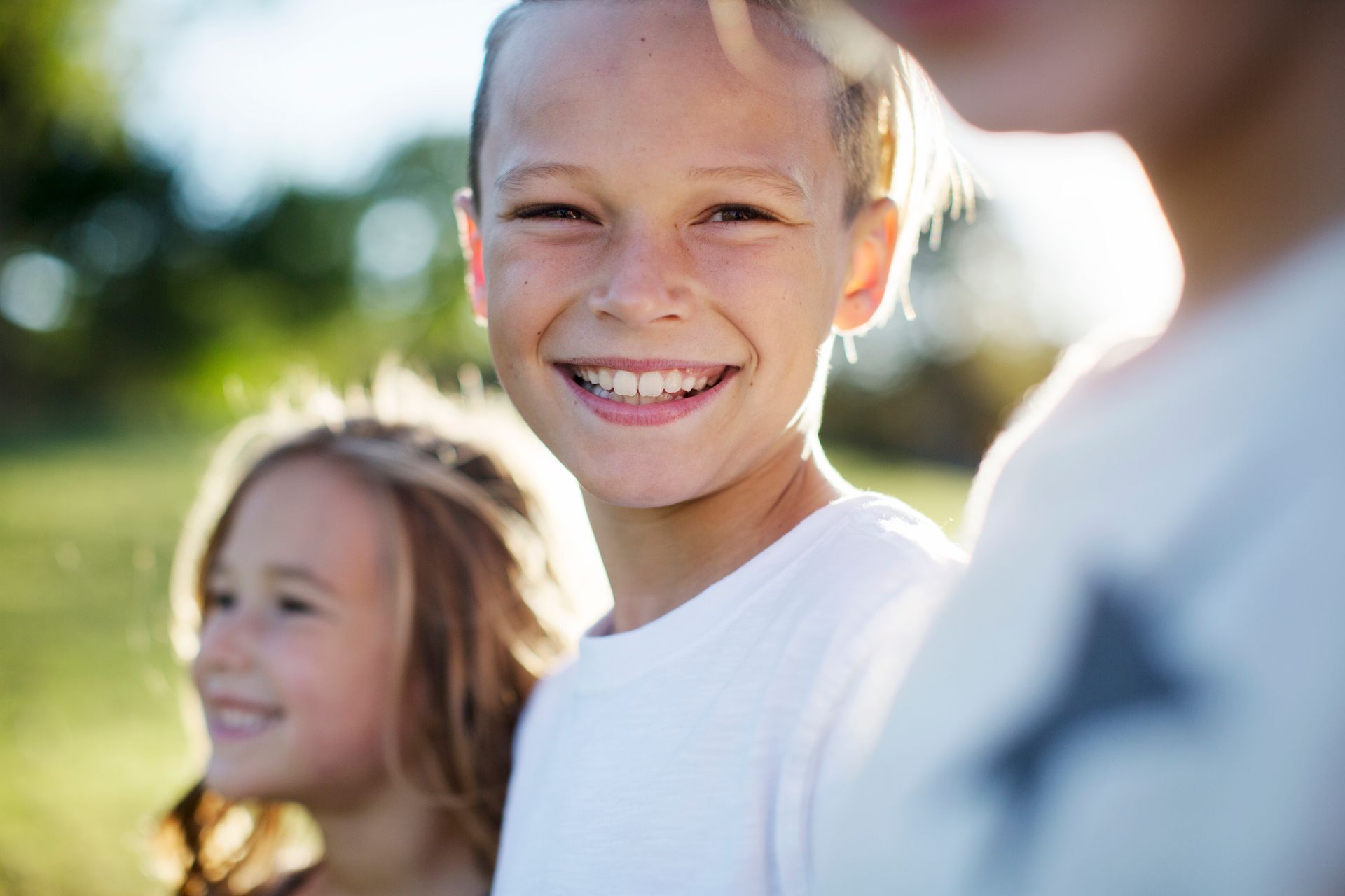 Smiling children outdoors in bright sunlight, with one child in a white shirt in the foreground