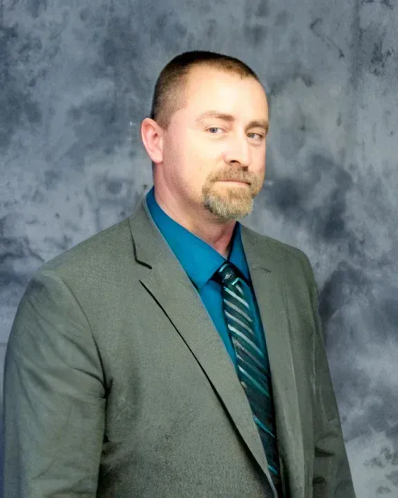 Portrait of a man in a gray suit with a teal shirt and striped tie against a mottled gray backdrop