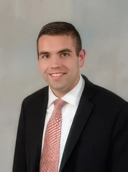 Headshot of a smiling person in a black suit, white shirt, and pink tie against a gray background