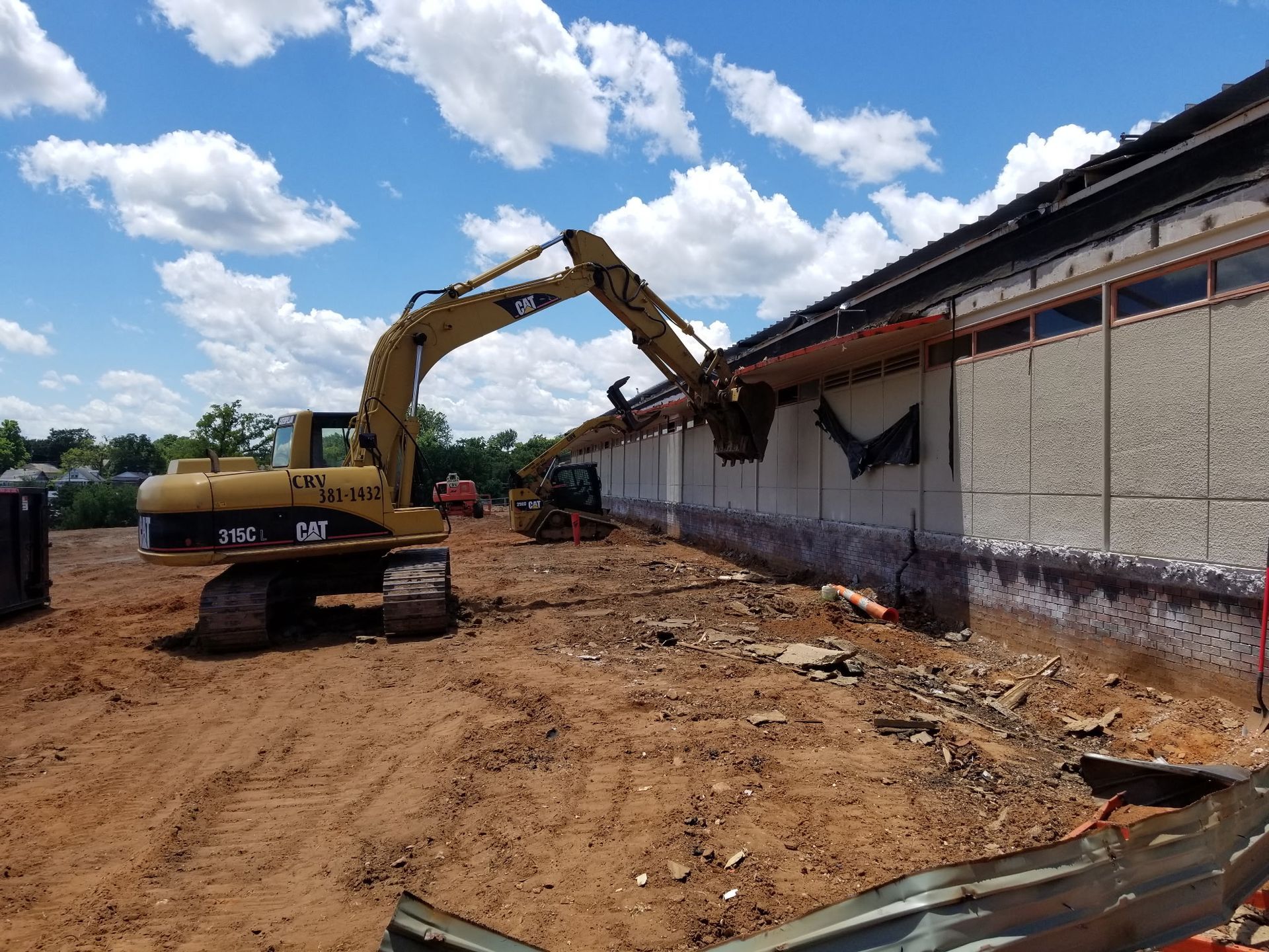 A yellow CAT excavator demoing the side of a building on a dirt construction site under a blue, cloudy sky.