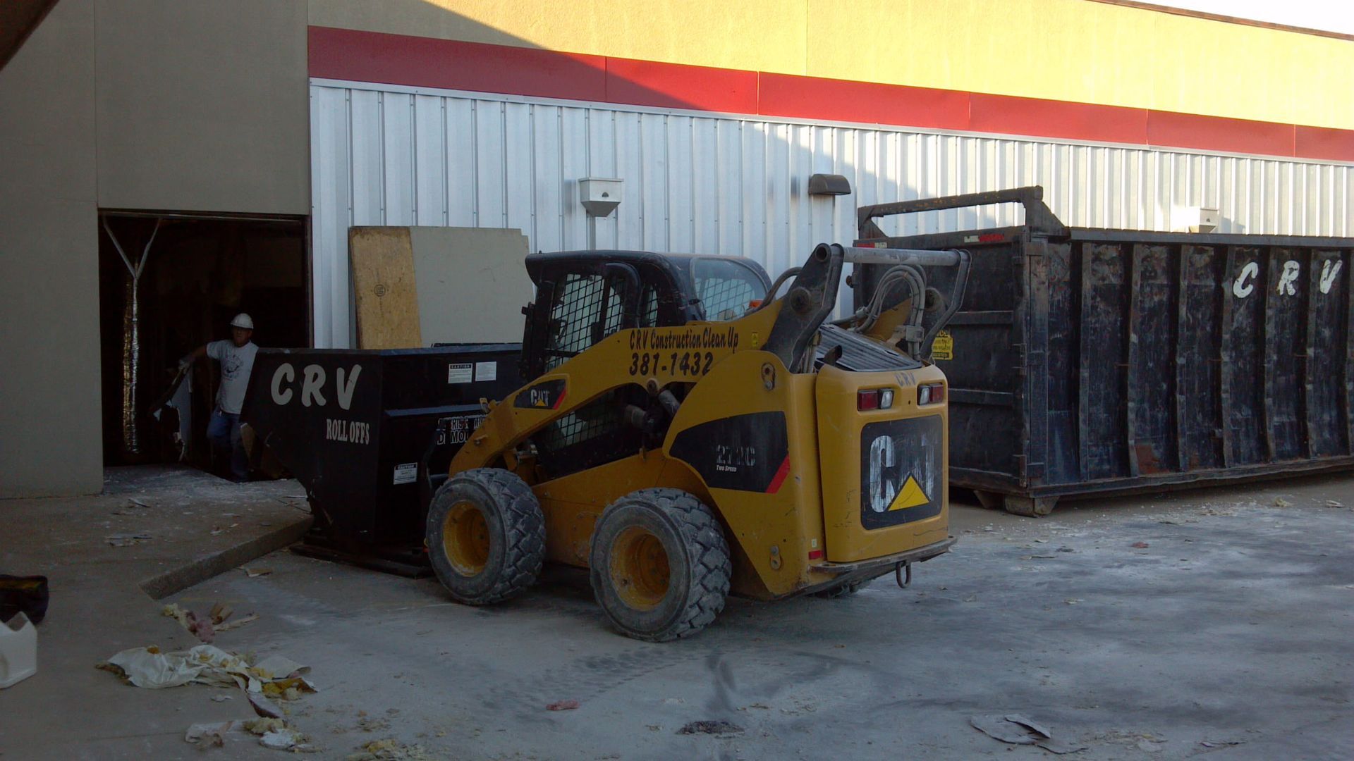 A yellow Caterpillar skid steer loads debris into a large black CRV dumpster next to a building.