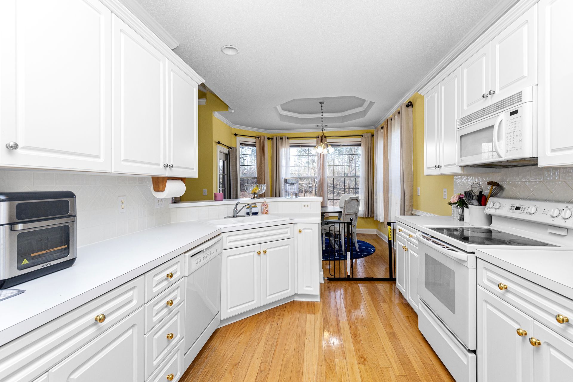 Bright white galley kitchen with wood floors, stainless appliances, and a small dining area at the far end