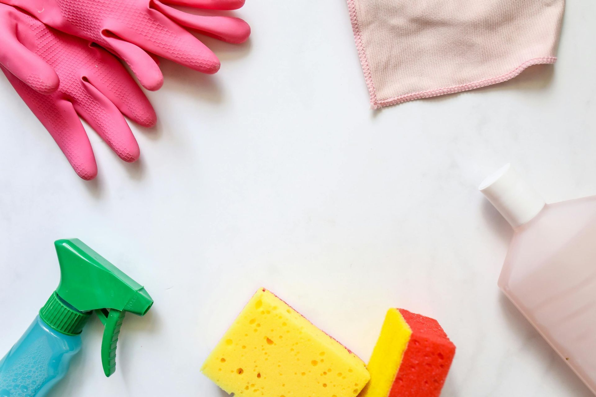 Cleaning supplies on a white surface: pink gloves, spray bottle, cloth, sponge, and disinfectant bottle.