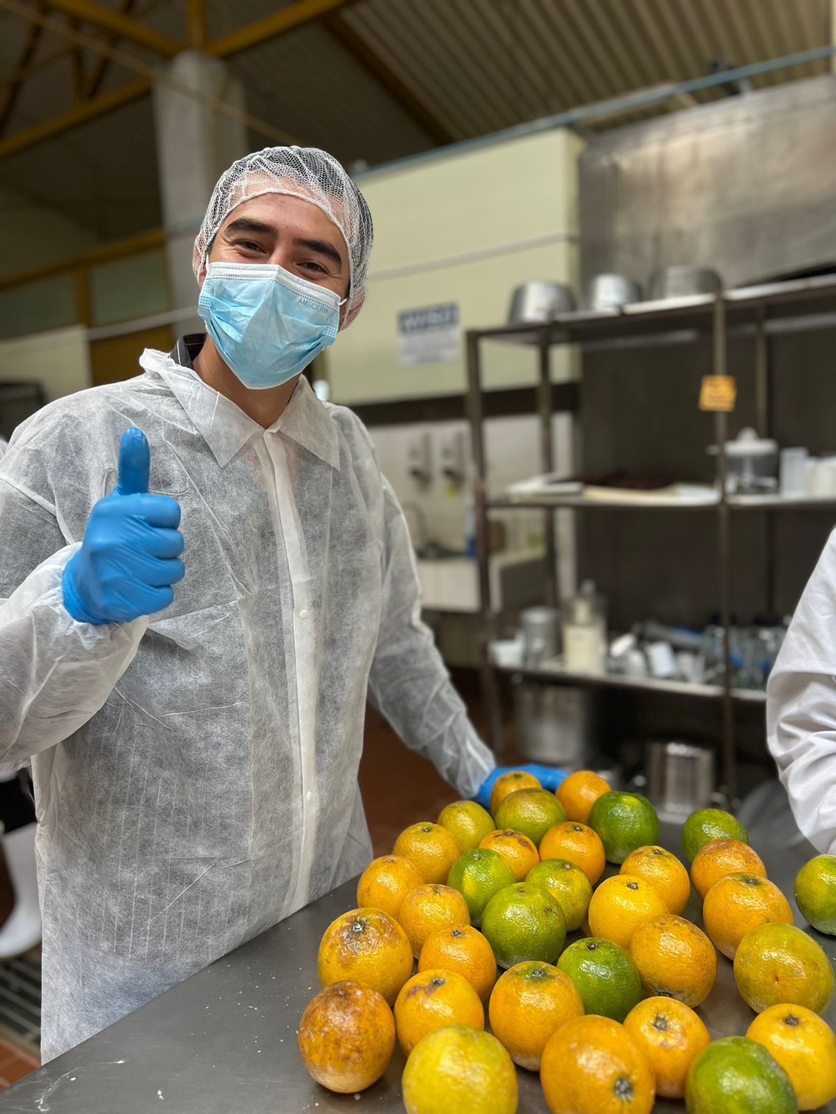 Un hombre que lleva una máscara y guantes hace un gesto de pulgar hacia arriba delante de una pila de naranjas.