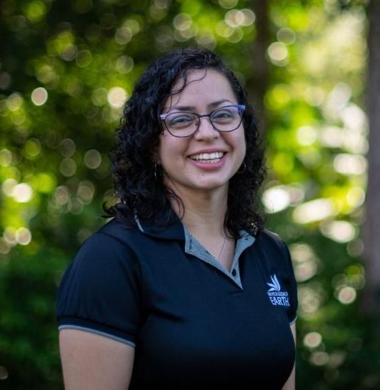 Mujer con gafas, camisa negra sonriendo al aire libre con fondo verde borroso.