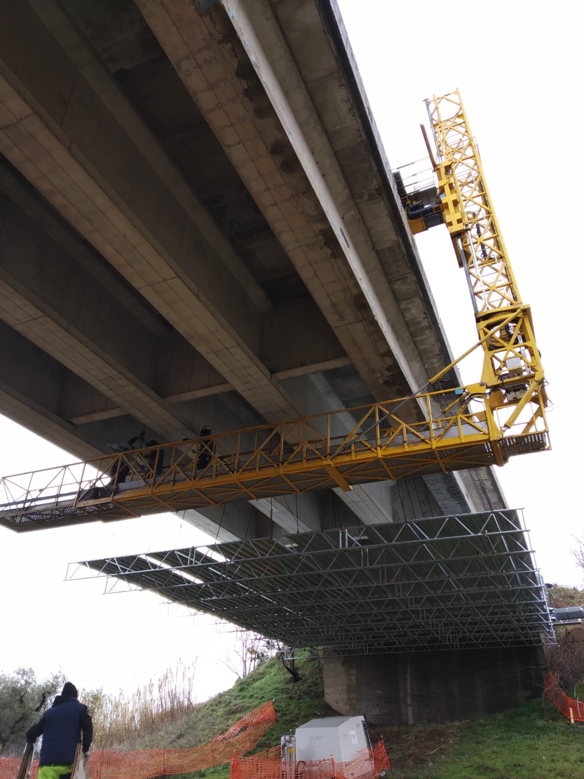 Operai su un'impalcatura sotto un ponte, con un braccio giallo di manutenzione proteso verso l'alto.