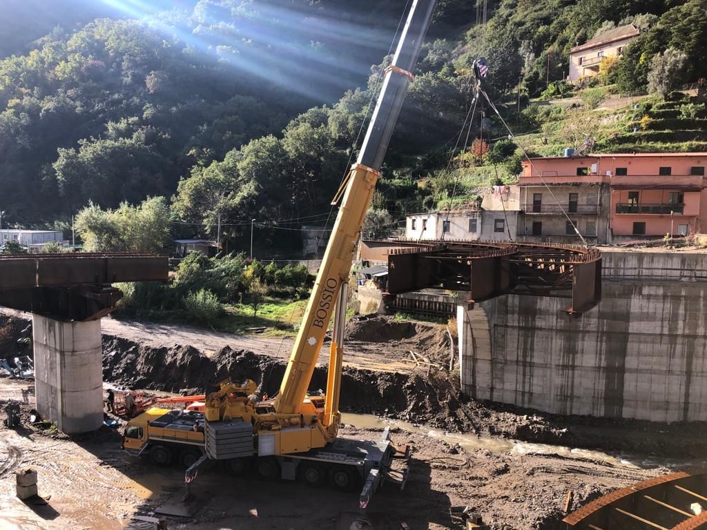 Una gru gialla solleva una sezione di ponte su un letto di fiume fangoso in un ambiente rurale con edifici e colline.