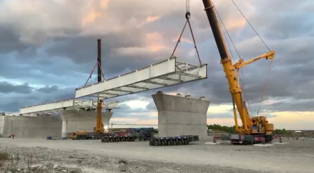 Gru che solleva una sezione di ponte in posizione durante la costruzione. Supporti in cemento grigio, cielo coperto.
