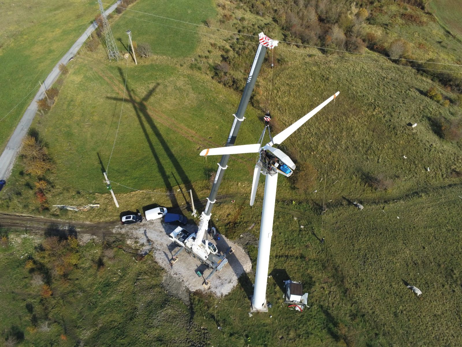 Vista aerea di una turbina eolica in costruzione su una collina erbosa; una gru solleva le pale.