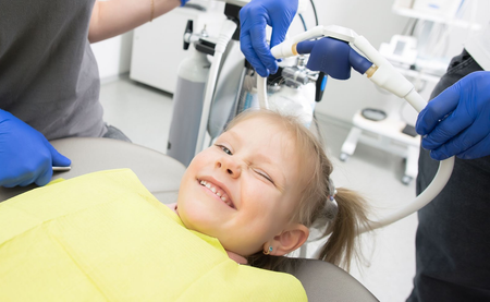 A dentist and assistant in blue scrubs wearing masks perform a checkup on a patient lying in a dental chair.