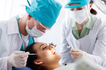 A dentist and assistant in blue scrubs wearing masks perform a checkup on a patient lying in a dental chair.
