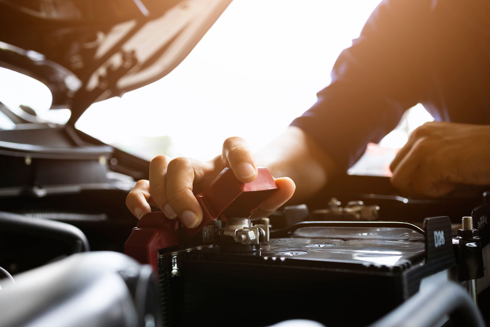 Person attaching a red battery clamp while working under the hood of a vehicle.