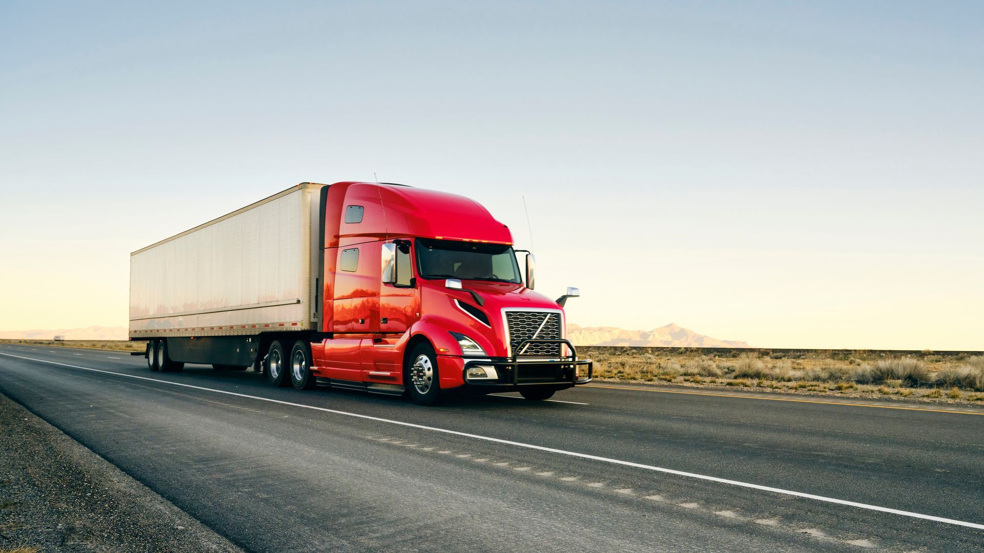 Red semi-truck driving on a highway, hauling a white trailer, desert landscape.