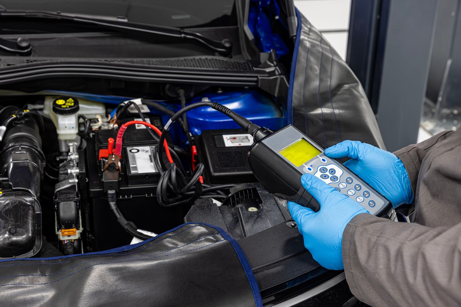 Mechanic tests a car battery with a diagnostic device, using blue gloves in an engine bay.