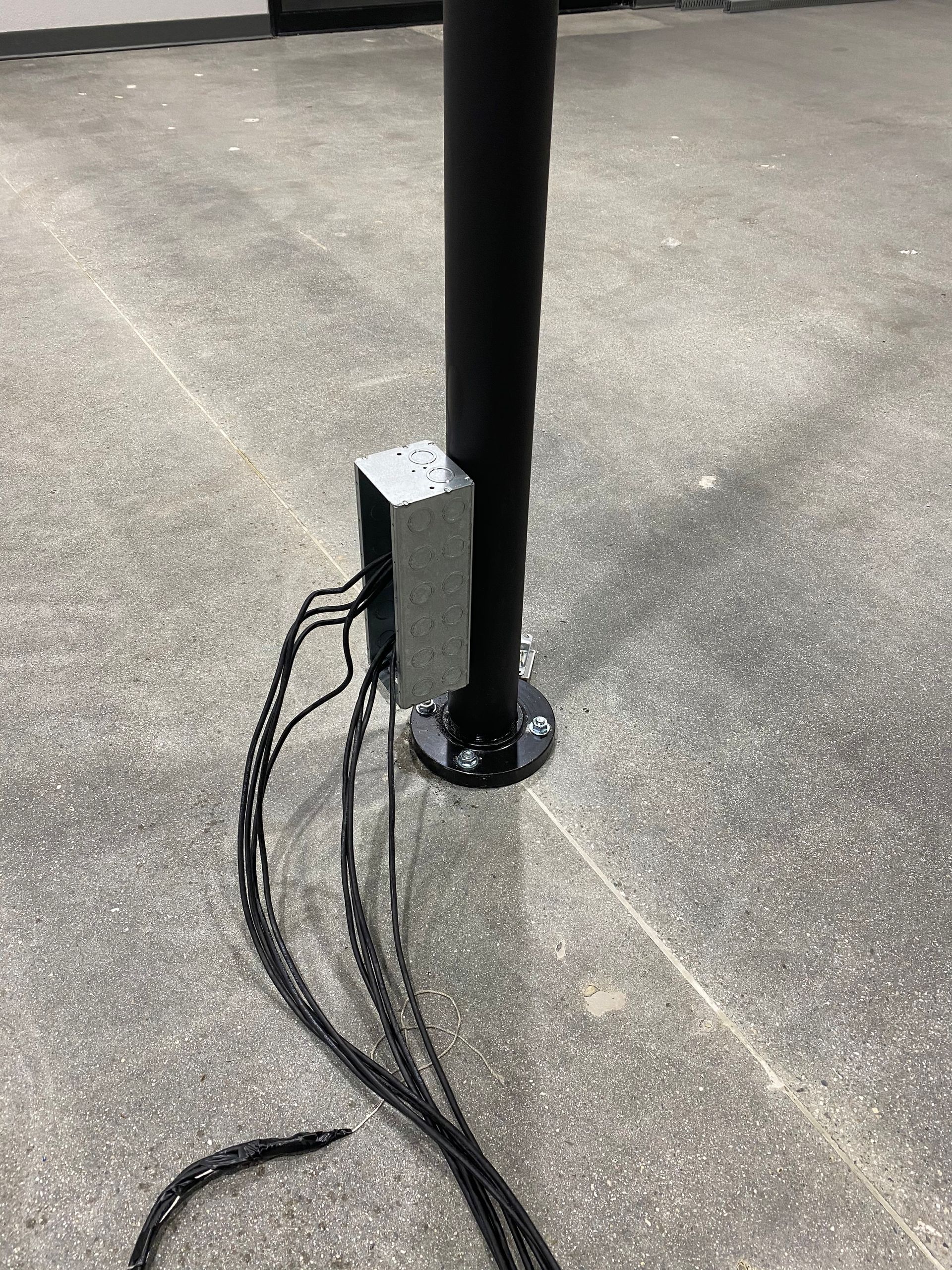 Black pole with a small box and loose cables on a concrete surface outdoors.