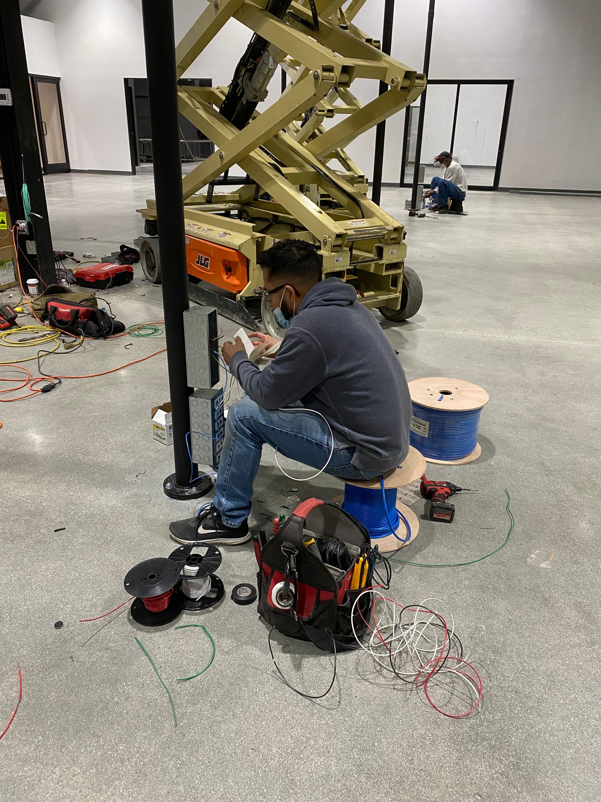 Technician crouched beside a scissor lift, working on tools and cables in a workshop.