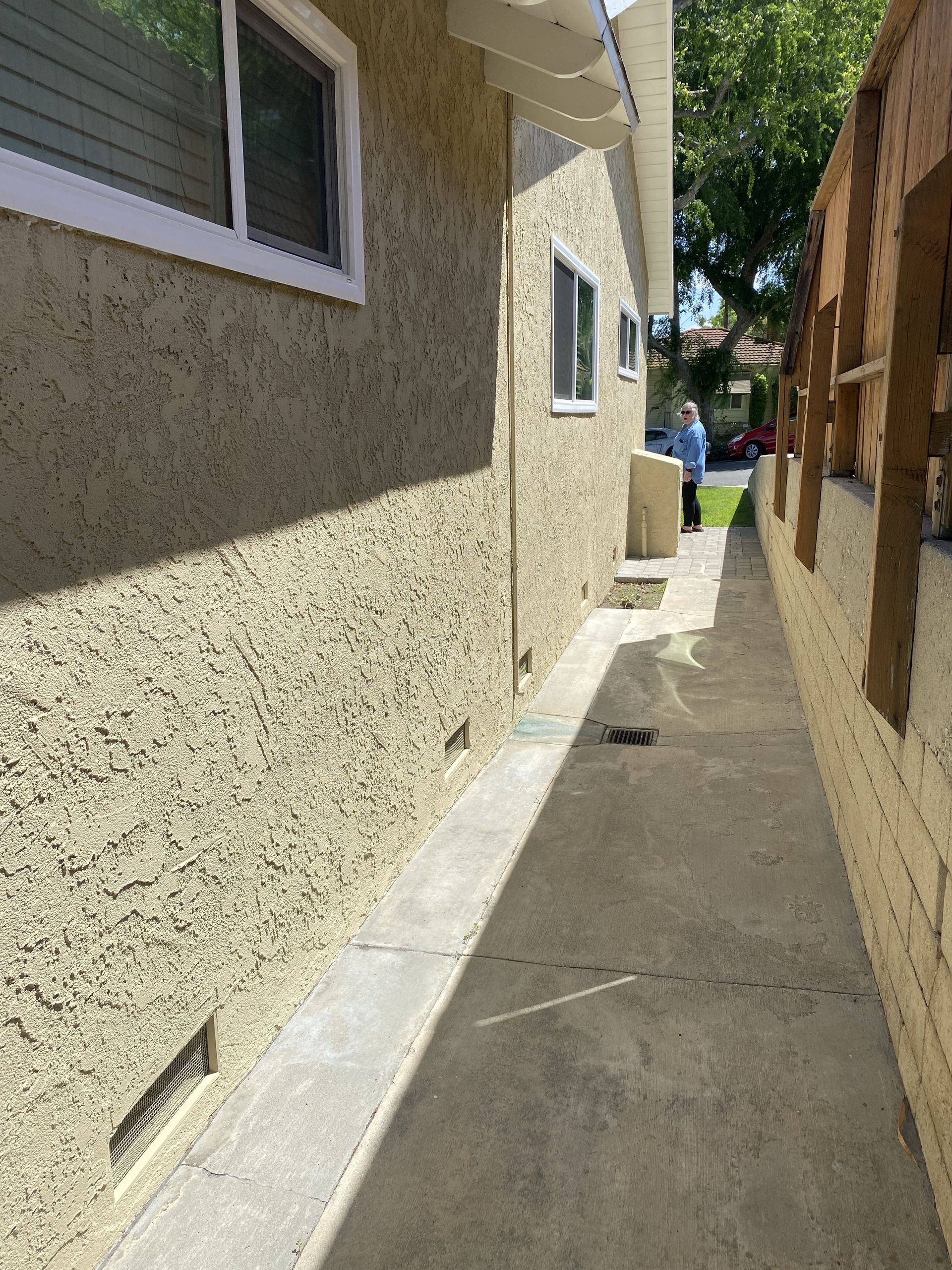 Narrow concrete walkway between a stucco house and wooden fence, leading to a driveway and blue trash bin.