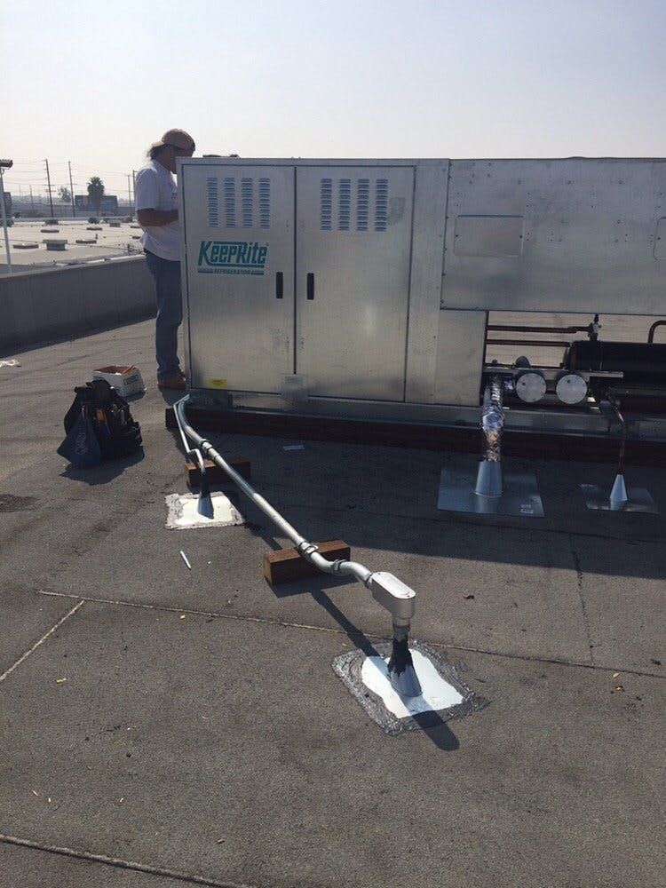 A technician stands next to an HVAC unit on a flat rooftop, inspecting a metal conduit connected to a sealed roof port.