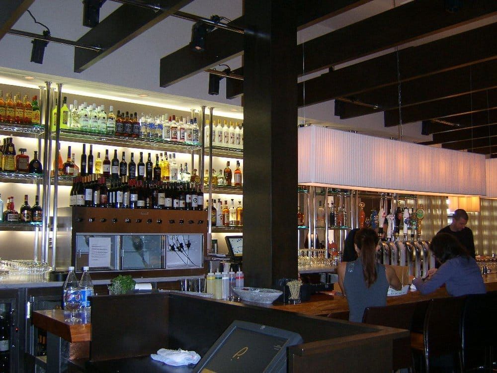 A modern bar with shelves of liquor bottles, exposed dark ceiling beams, and customers sitting at a wooden counter.
