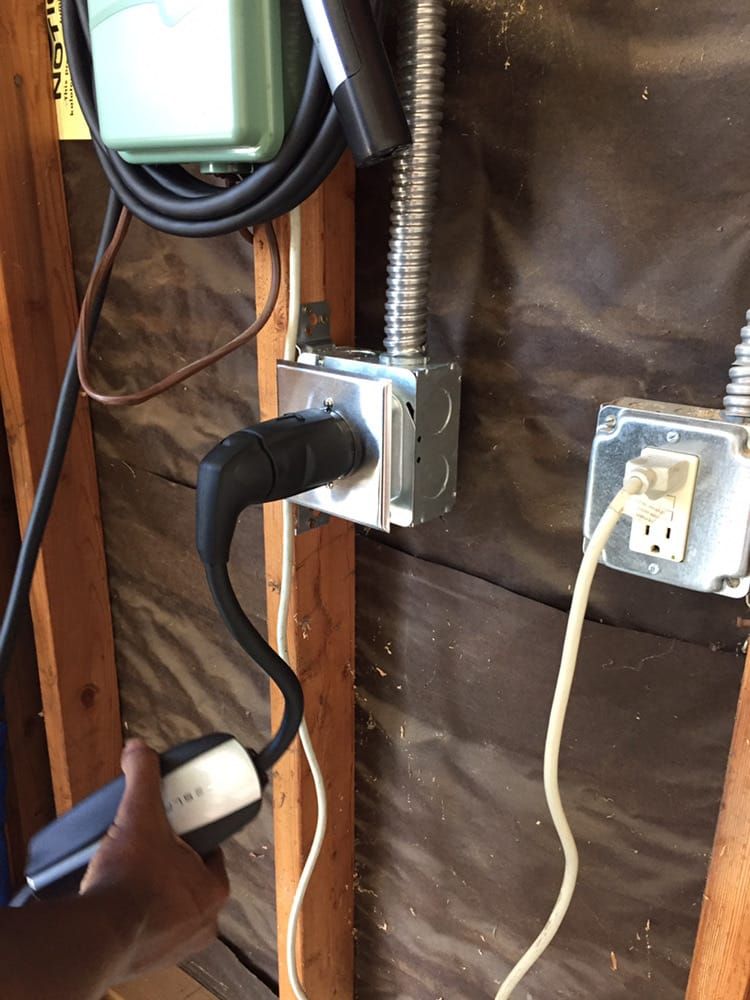 A hand plugs an electric vehicle charger into a wall-mounted metal outlet box in a garage.
