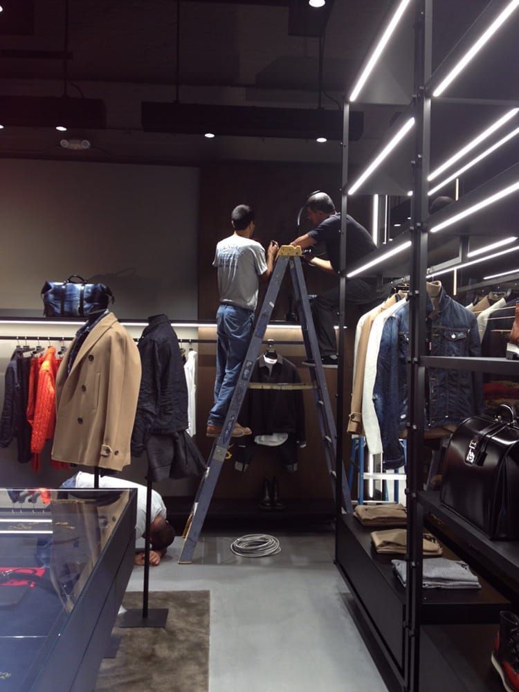 Two workers on a ladder installing lighting in a clothing store with displays of apparel and accessories.