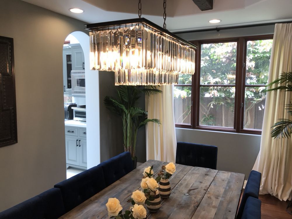 A modern dining room featuring a rectangular crystal chandelier above a wooden table, blue chairs, and off-white curtains.