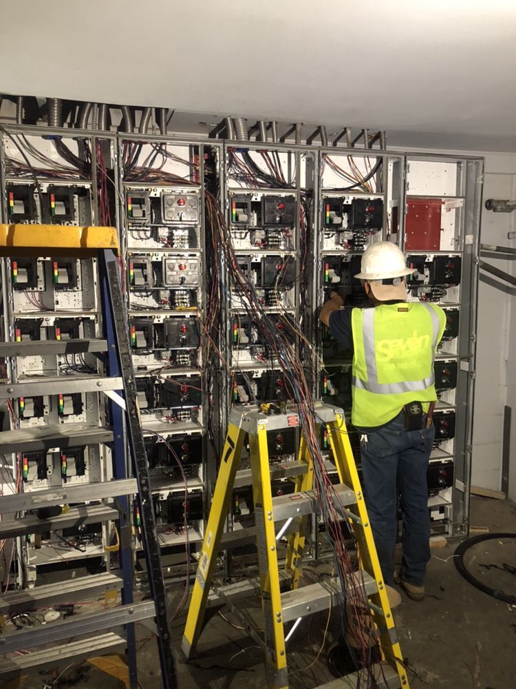 Electrician in a high-visibility vest and hard hat works on wiring inside a large, multi-section electrical cabinet.