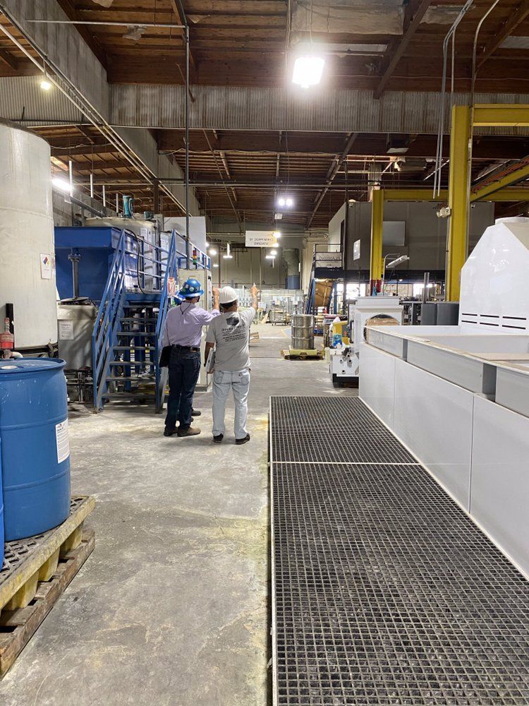 Two workers in hard hats and safety gear stand in an industrial facility, observing equipment on a factory floor.