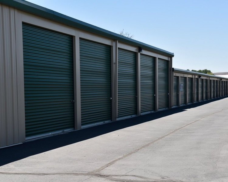 Row of green storage unit doors along a long concrete driveway under a clear sky