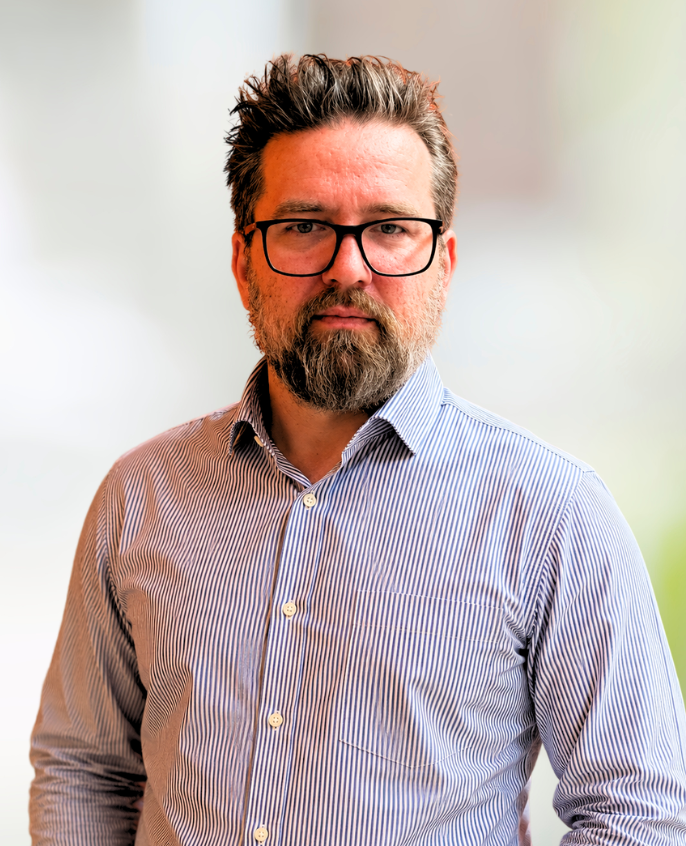 Man wearing glasses and a striped button-down shirt, standing against a softly blurred background.