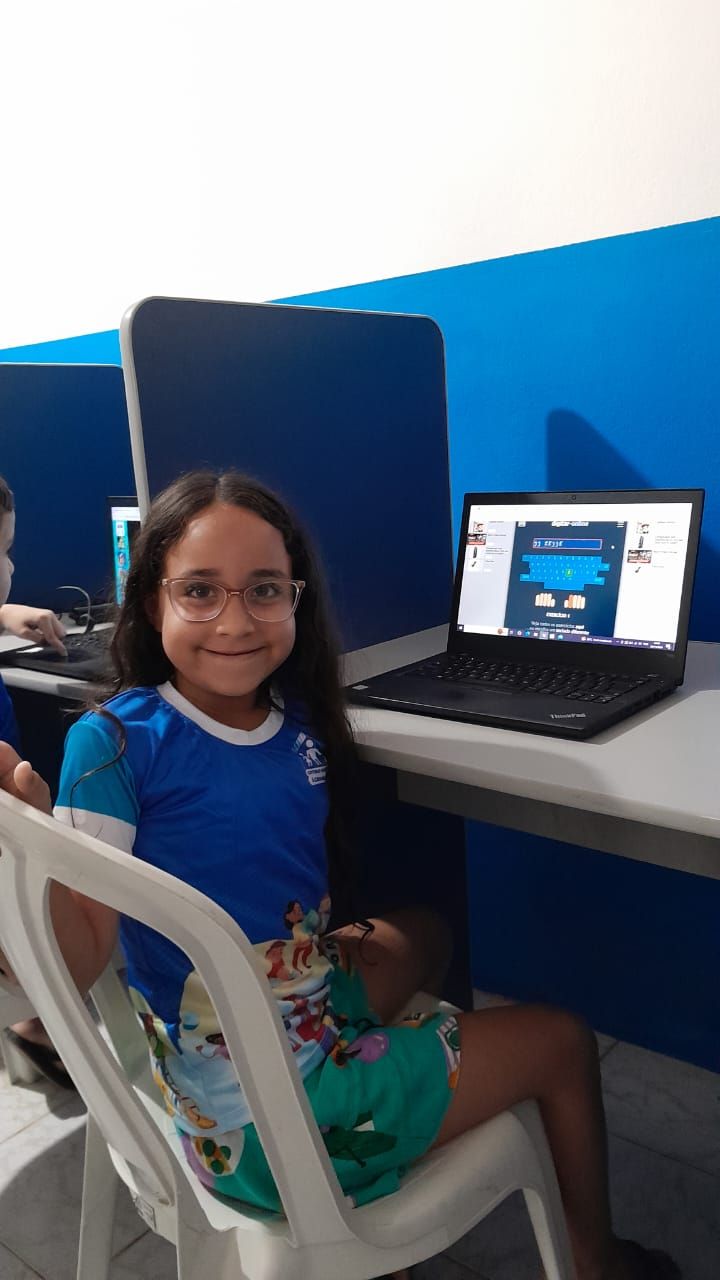 Uma pessoa jovem, de cabelos compridos e óculos, está sentada em uma mesa, sorrindo ao lado de um laptop