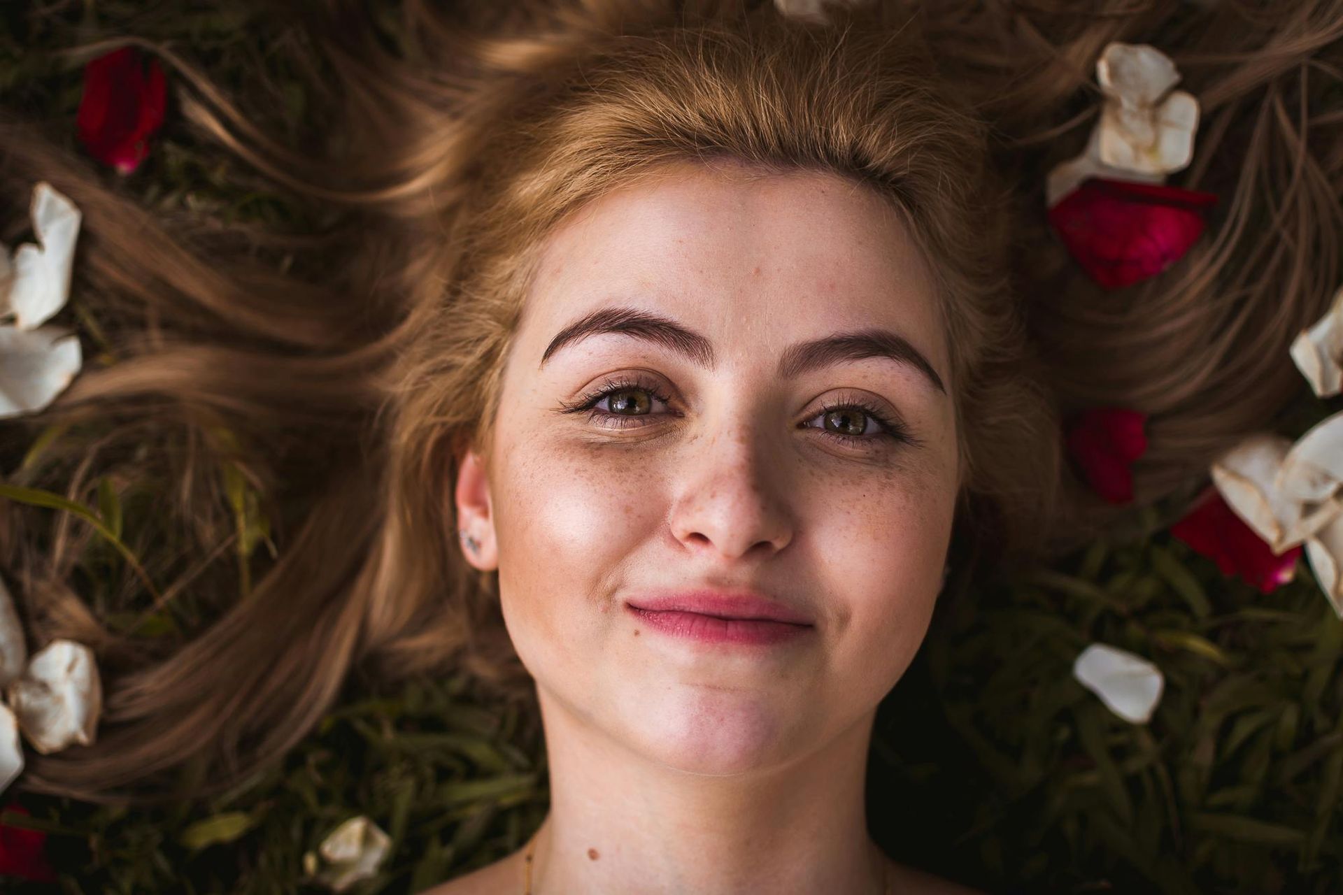 A person with long blonde hair lying on grass surrounded by scattered white and red rose petals, smiling at the camera.