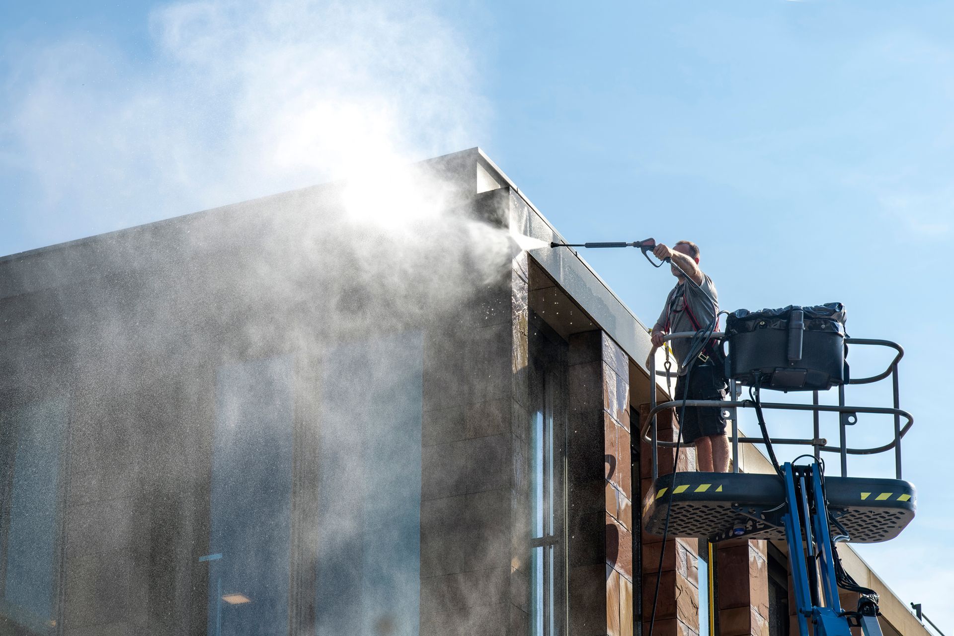 Worker pressure-washing the side of a building from a lift, with water spraying in the sunlight