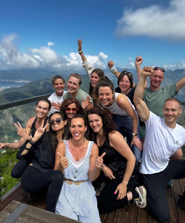 Een groep mensen poseert vrolijk buiten met een bergachtig landschap op de achtergrond. Duimen omhoog, armen geheven, zonnige, blauwe lucht.
