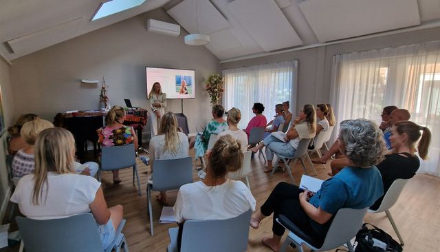 Een presentatie in een lichte ruimte; een vrouw spreekt voor een zittend publiek.
