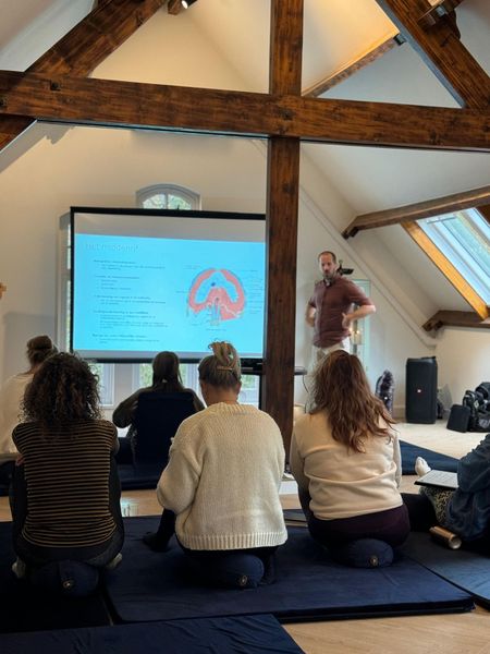 Een man presenteert een dia aan een groep mensen die zitten. Ze bevinden zich in een kamer met houten balken en een projector.