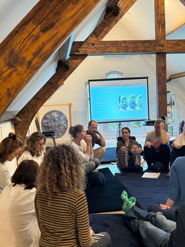 Mensen zitten in een kring tijdens een presentatie, op een blauw tapijt, in een ruimte met houten balken en een scherm.