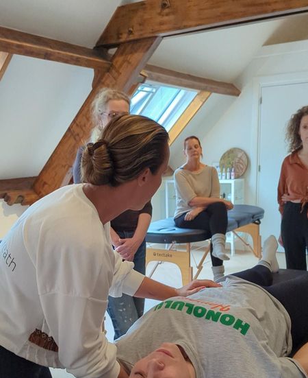 Een vrouw demonstreert een techniek op een persoon die op een massagetafel ligt, terwijl anderen toekijken in een kamer met een dakraam.