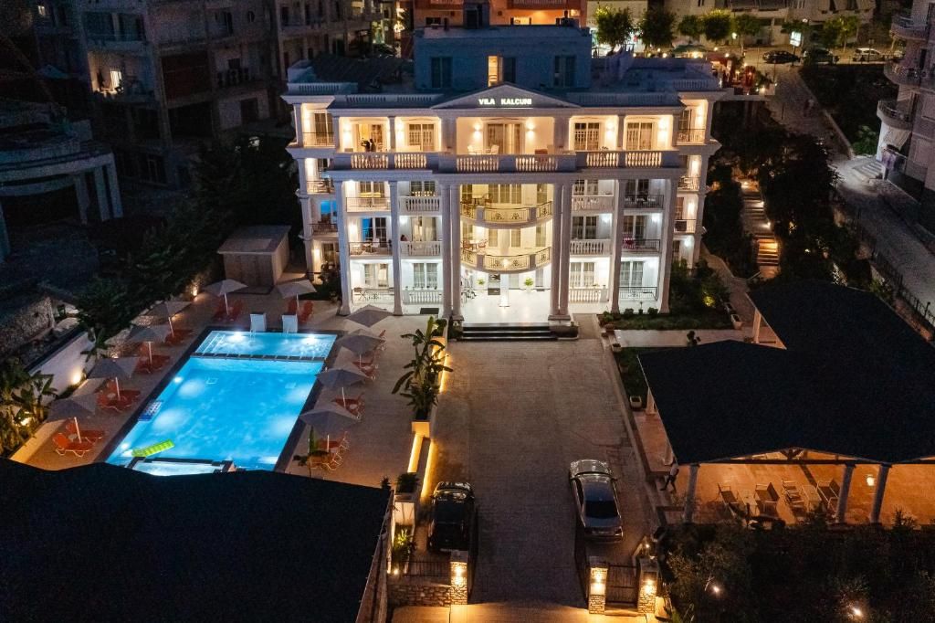 Vedere panoramică nocturnă asupra Vila Kalkuni Luxury Resort, cu luminile, piscina, șezlongurile și foișorul vizibile în cadru. Atmosfera nocturnă scoate în evidență ambianța relaxantă a resortului din Sarandë.