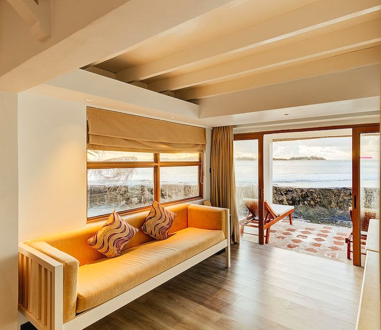Living luminos cu o canapea și pereți galbeni la Cinnamon Dhonveli Maldive, cu ieșire directă spre mare. Spațiul deschis oferă o priveliște directă asupra apelor turcoaz din jurul resortului.