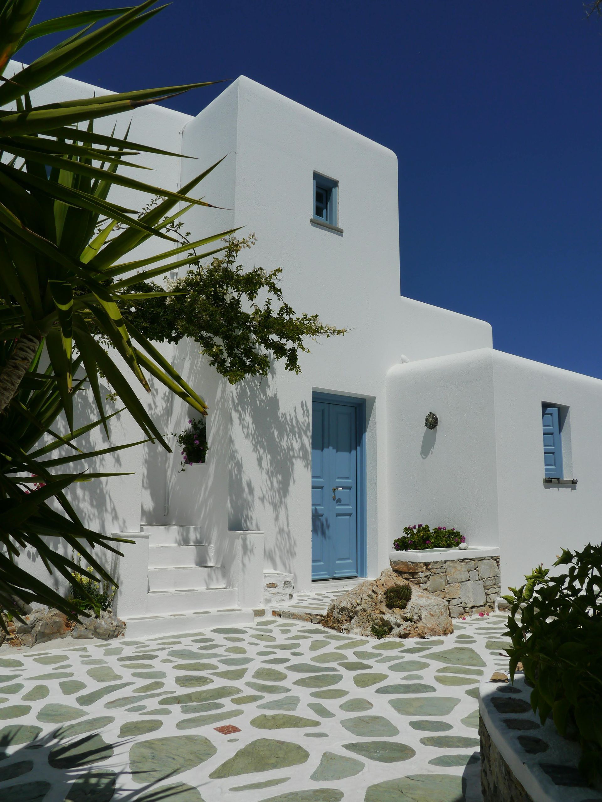 Intrarea vilei de vacanță din Creta, Grecia. Vila este joasă, văruită în alb, cu ferestre mici și albastre, în stil tradițional grecesc.
