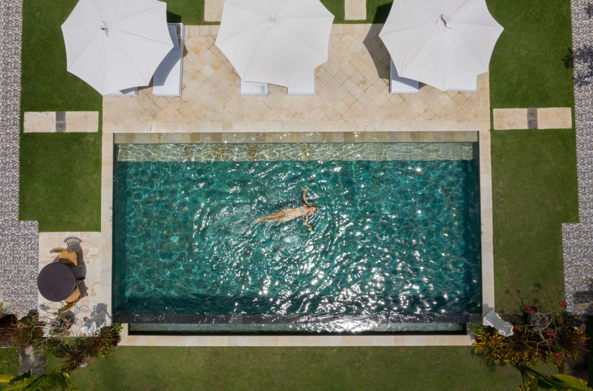 Imagine de sus de la o vilă de vacanță cu o femeie plutind pe spate într-o piscină la soare, cu umbreluțe lângă. 