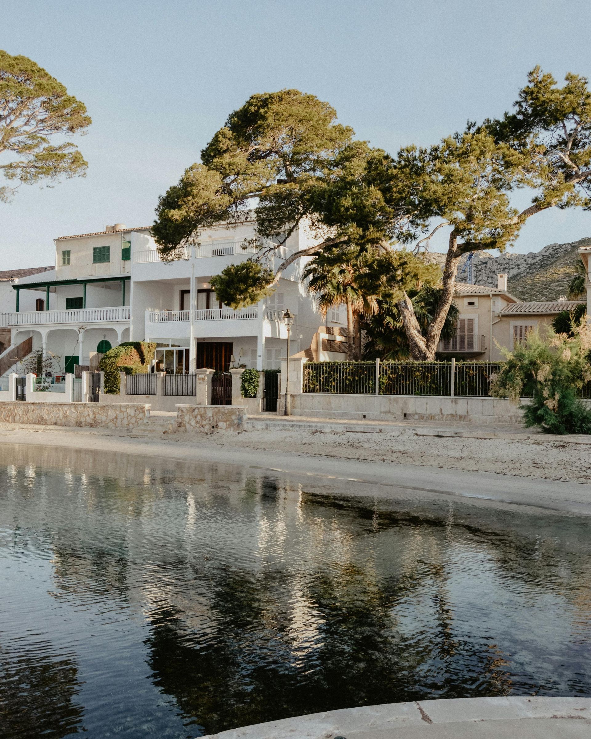 O vilă de vacanță din Palma de Mallorca. În prim-plan este chiar marea, iar apoi se observă vila albă și grădina cu vegetație mediteraneană.