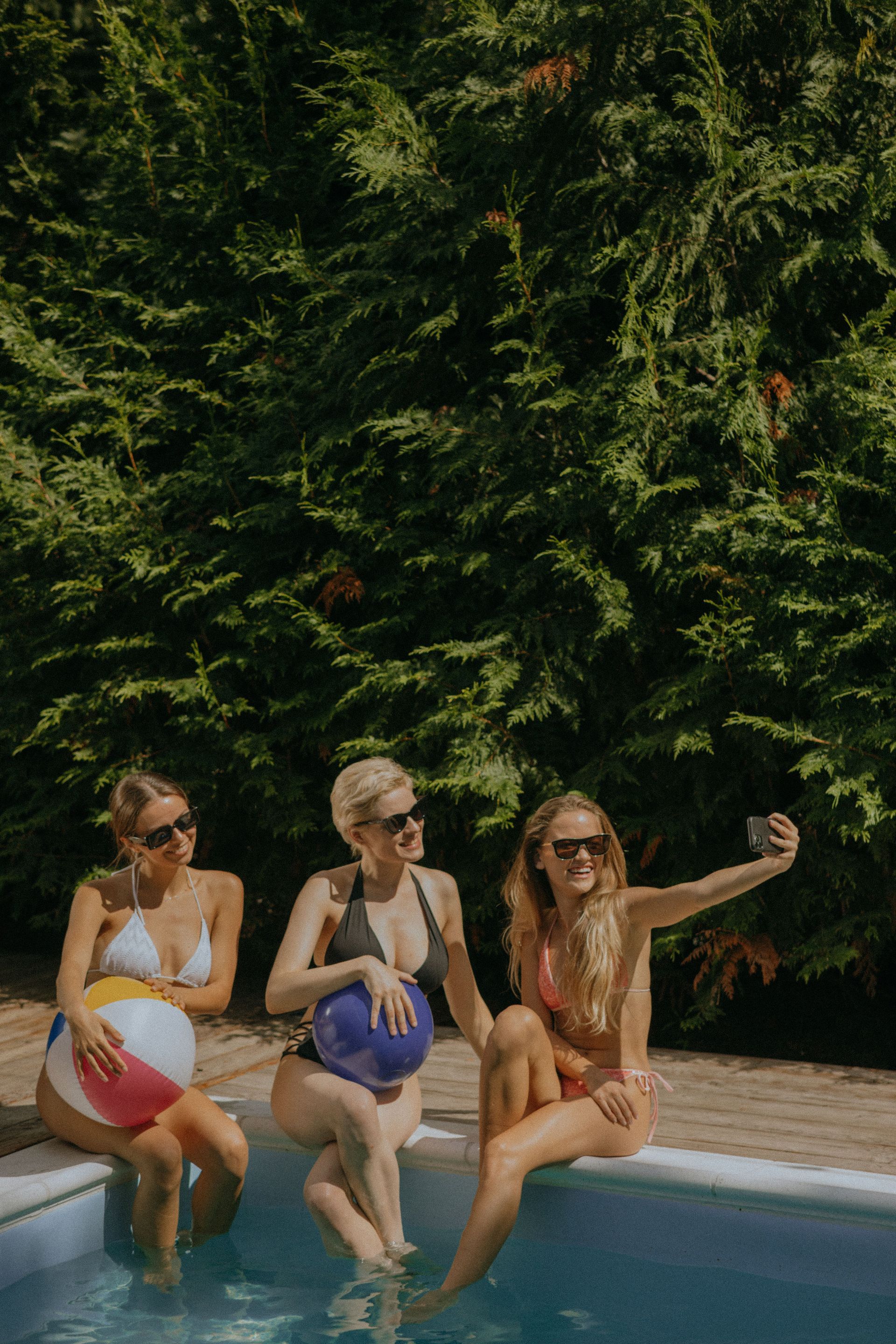 Trei fete râzând și făcându-și un selfie pe marginea piscinei de la vila de vacanță rezervată, cu picioarele în apă.