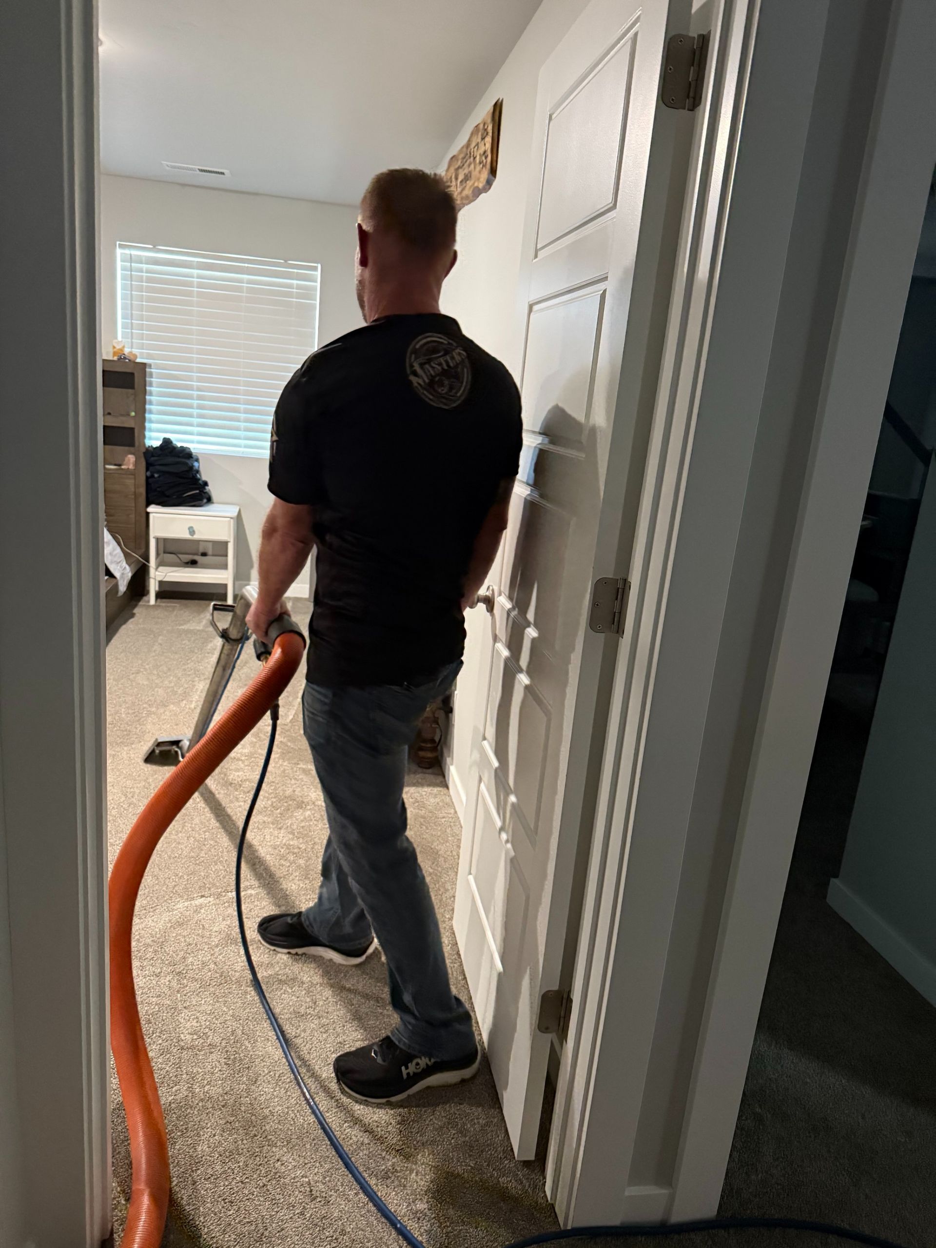 Person using an orange vacuum hose in a carpeted hallway beside a white door.