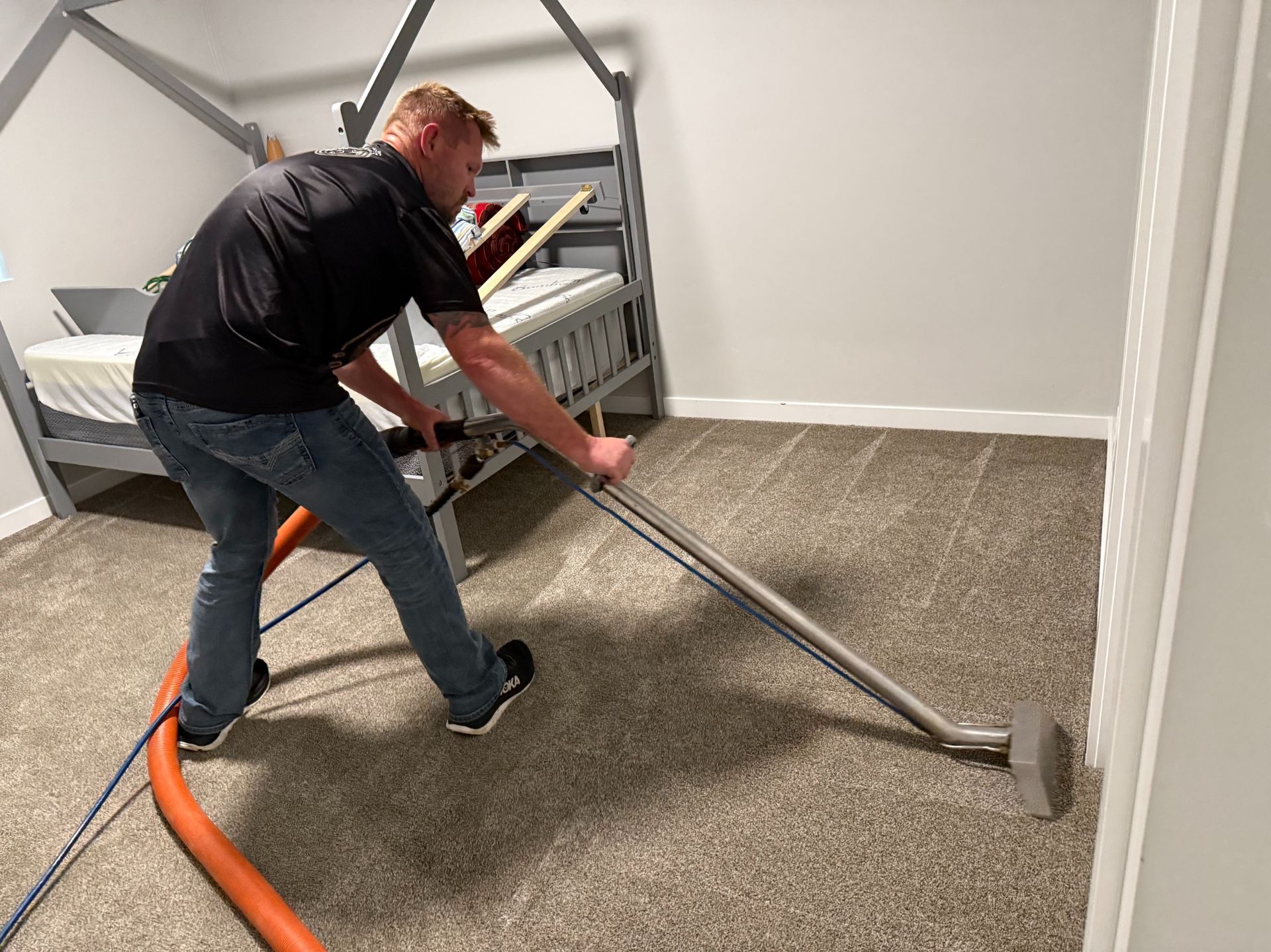 Man vacuuming beige carpet in a small room beside a metal bunk bed frame.