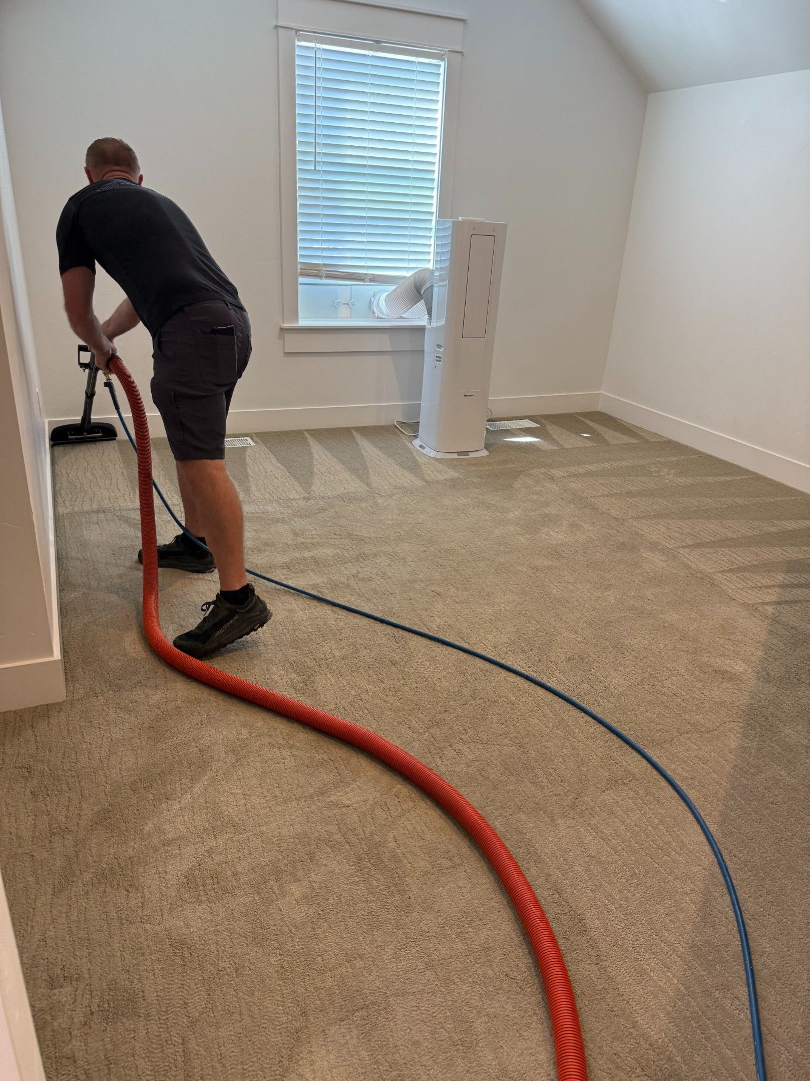 Worker vacuuming carpet in a small empty room with hoses on the floor.