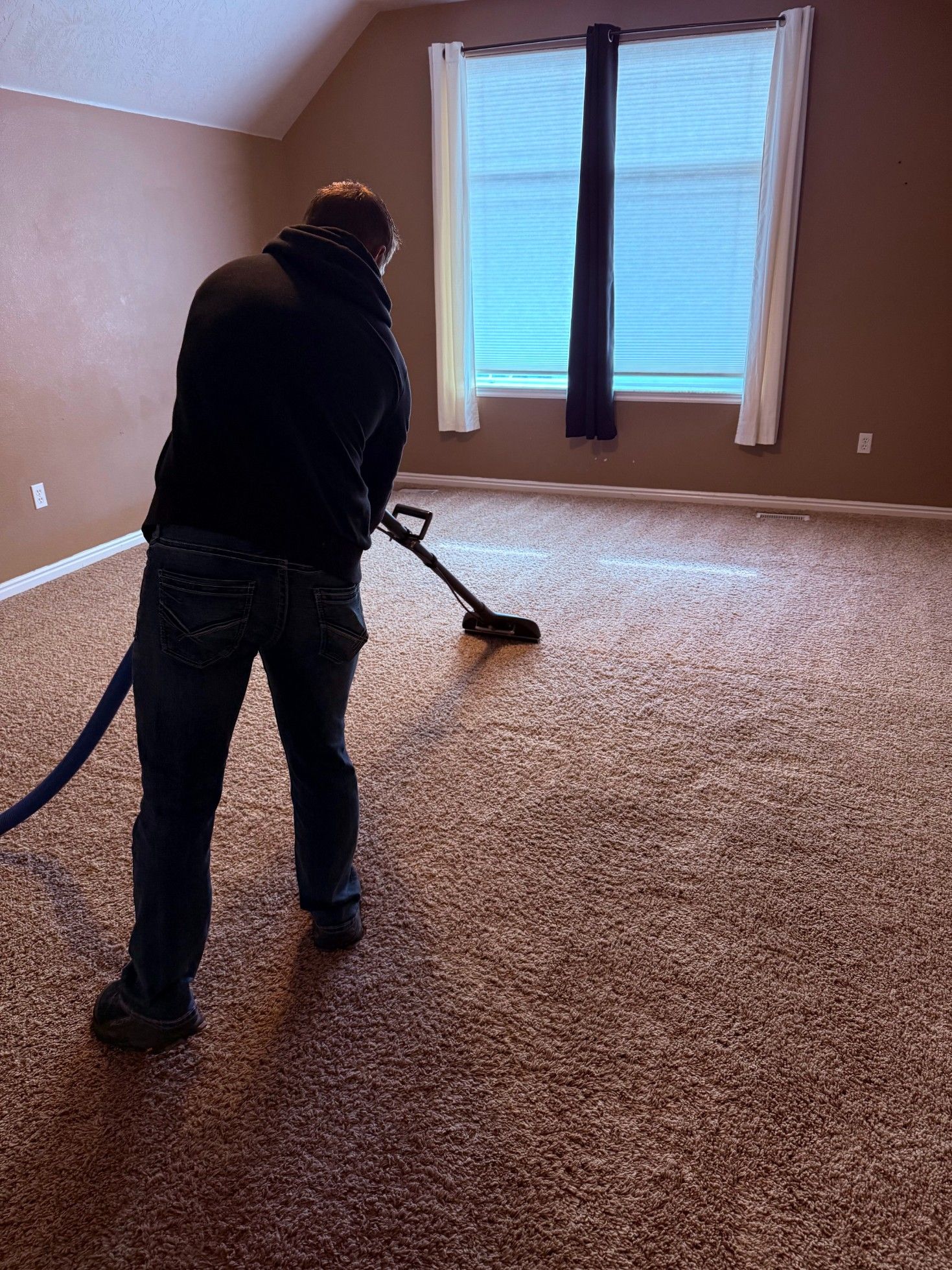 Person vacuuming a carpeted room with beige walls and a large window.