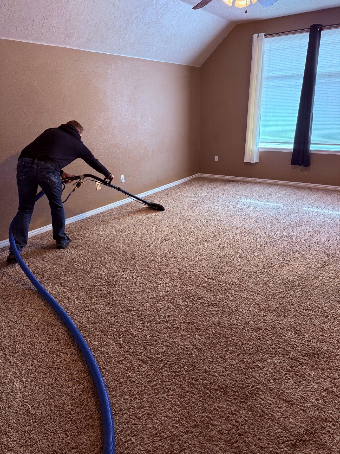 Person vacuuming beige carpet in an empty room with tan walls and a window with blinds