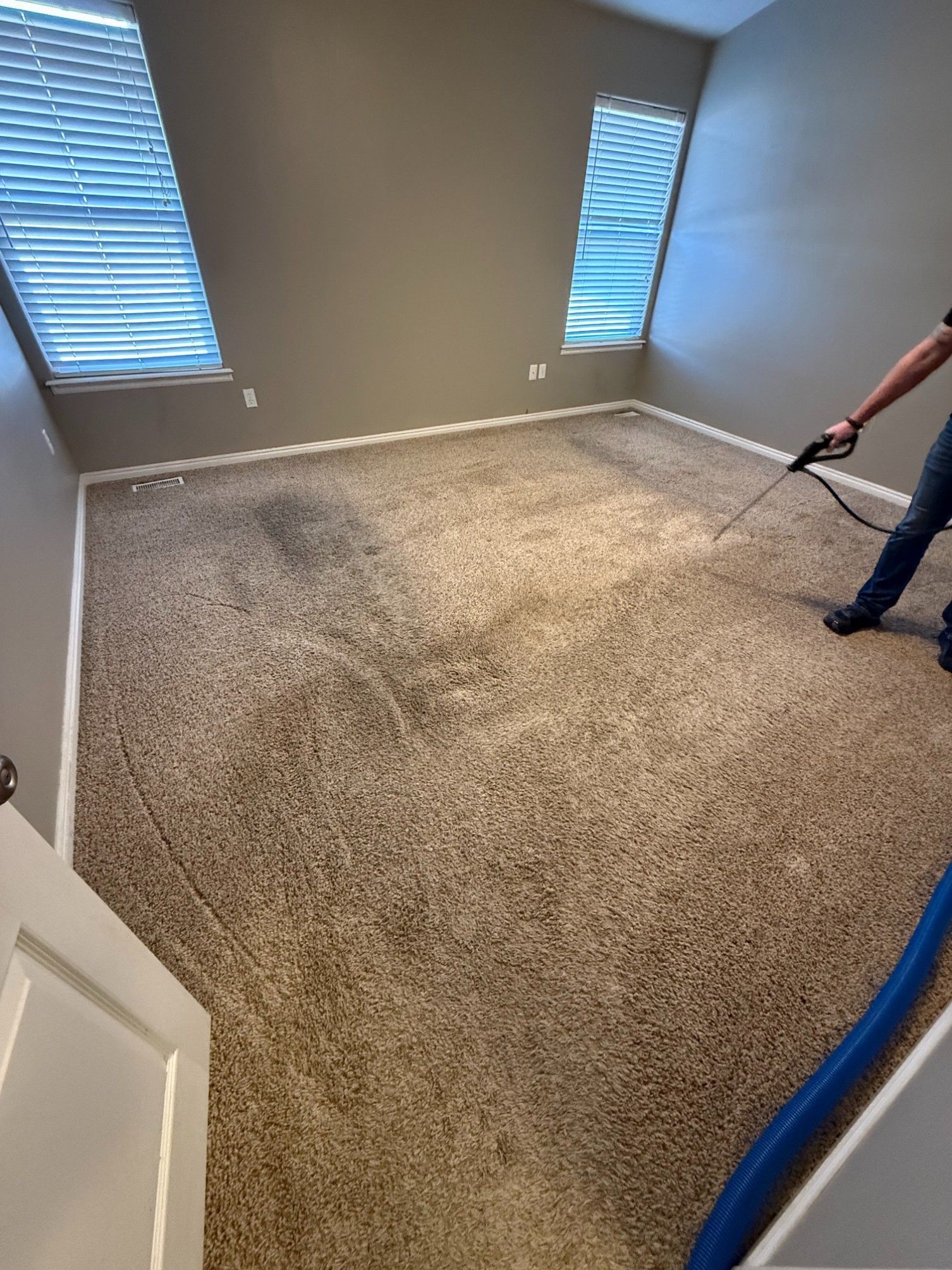 Carpet cleaner spraying beige carpet in an empty room with gray walls and windows with blinds